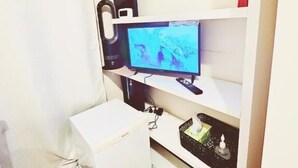 Une pièce comprenant un bureau blanc, un ordinateur, un téléviseur et une étagère sur laquelle sont disposés divers objets.