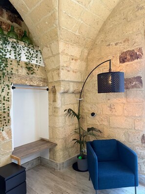 Habitació amb paret de pedra, una cadira blava, una làmpada de peu, una planta i una banca de fusta.