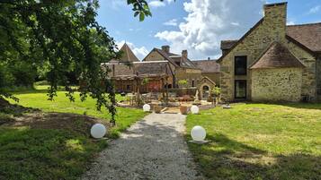 A rustic courtyard with a gravel path leading to a wooden pergola, stone buildings, and a clear sky.