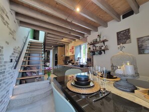 Una cocina con vigas de madera, una escalera, un comedor con una mesa dispuesta para dos personas y vistas al salón.