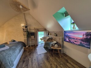 Un dormitorio con zona de descanso en altura, suelo de madera, escritorio con silla, lavamanos y un ventanal grande.