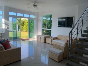 A spacious living area with a large sliding glass door leading to a garden, a ceiling fan, a flat-screen TV mounted on the wall, and a staircase with metal railings. - Serene Seaside Retreat in Juan de Acosta - Private Pool & Comfort (Juan de Acosta)
