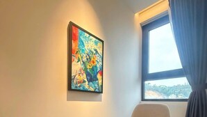 A room with a beige armchair, a small round table with a cup and saucer, and a colorful painting on the wall. - Cozy studio in Sapphire Ha Long with Sun Wheel view (Hồng Gai)