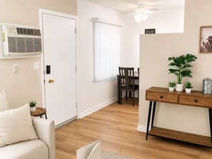 A room with a sofa, a dining table with chairs, a wooden desk with plants, and an air conditioning unit. - Hidden NoHo Nook (Los Angeles)