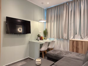 Een moderne woonkamer met een flatscreen televisie, een eethoek met een tafel en stoelen, een grijze bank en een groot raam met gordijnen.