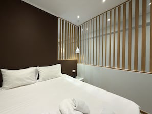 Een hotelkamer met een groot bed, een nachtkastje, een lamp en een raam met verticale jaloezieën.