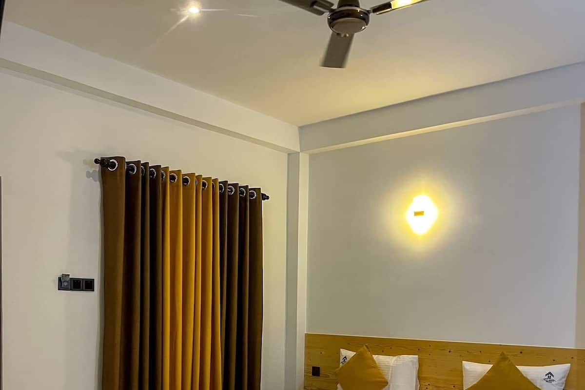 A hotel room with a bed, a ceiling fan, a wall-mounted light, and a window with curtains.