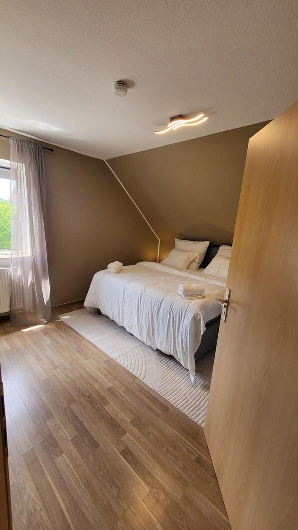 Ein Schlafzimmer mit Holzboden, einem Bett mit weiĂer BettwĂ€sche, einem Fenster mit leichtem Vorhang und einer Deckenleuchte.