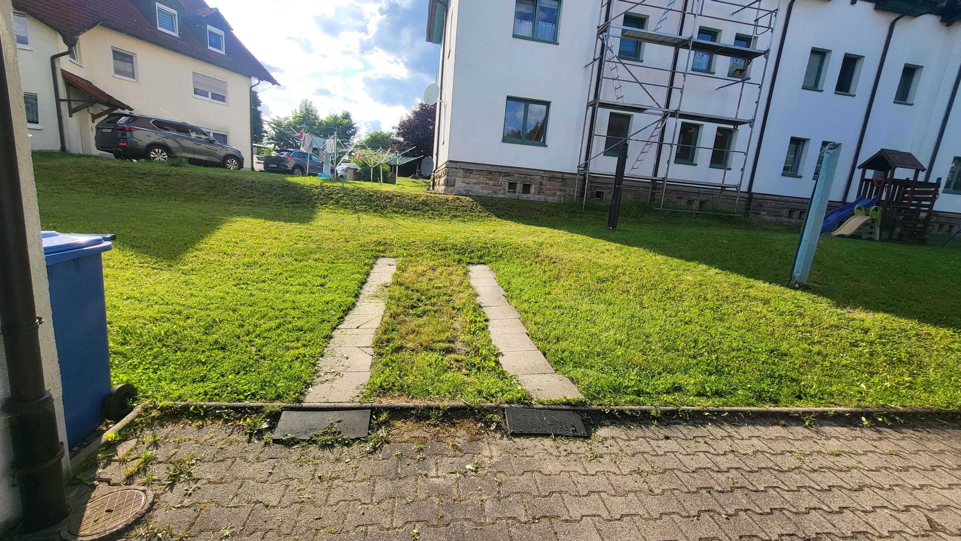 A paved pathway leading uphill with a grassy area, a parked car, and a building with multiple windows and scaffolding.