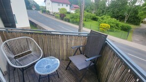 Un balcon avec deux fauteuils en osier et une petite table, donnant sur une route et sur un espace verdoyant.
