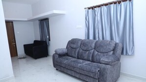 En stue med en grå sofa, en mørk lænestol, et hvidt loft og et vindue med lyseblå gardiner.