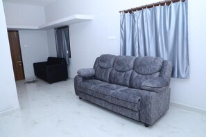 A living room with a grey sofa, a dark armchair, a white ceiling, and a window with light blue curtains. - Shreesree Homestay (Tiruvannamalai)