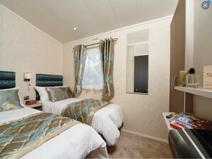 A hotel room with two beds, a window with curtains, a bedside table with a lamp, and a small shelf with items. - Ocean Edge Leisure Park - Holiday Accommodation 1317 (Morecambe)