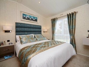 A hotel room with a bed, bedside tables, a headboard, a wall-mounted artwork, and a window with curtains. - Ocean Edge Leisure Park - Holiday Accommodation 1317 (Morecambe)