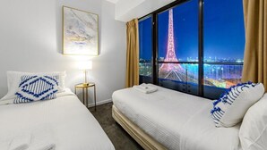 A hotel room with two beds, a night view of the Eiffel Tower, a framed artwork, and a bedside table with a lamp. - Exclusive Stays - Gallery Towers (Southbank)