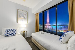 A hotel room with two beds, a night view of the Eiffel Tower, a framed artwork, and a bedside table with a lamp. - Exclusive Stays - Gallery Towers (Southbank)
