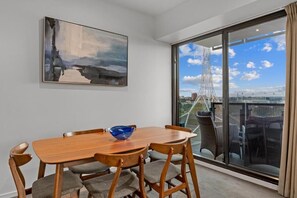 A dining area with a wooden table and chairs, a blue bowl, a large abstract painting, and a view of a cityscape through a glass door. - Exclusive Stays - Gallery Towers (Southbank)