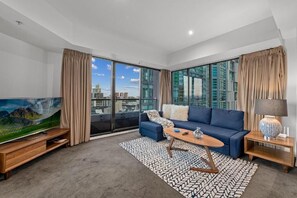A modern living room with a large TV, a blue sofa, a coffee table, and a view of the city through large windows. - Exclusive Stays - Gallery Towers (Southbank)