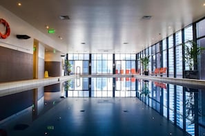 Uma piscina interior moderna com paredes de vidro transparente, sinalização de saída e áreas de estar.