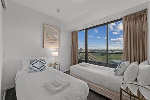 Quarto de hotel com duas camas, uma janela grande com vista para a cidade, um quadro na parede e uma pequena mesa com uma luminária.