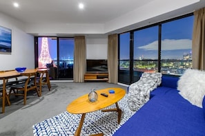 Habitación de hotel moderna con un ventanal grande que ofrece vistas a la ciudad, un sofá con una manta suave, una mesa de centro de madera y una zona de comedor con mesa y sillas.