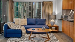 A modern living room with a sofa, coffee table, and lamp, a kitchen area with cabinets and appliances, and a large window with a city view. - Exclusive Stays - Gallery Towers (Southbank)
