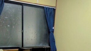 Una cama individual con colchón, almohadas y manta. Se ve una ventana con cortinas, una mesita de noche con lámpara e un enchufe de pared.