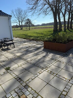 Een geplaveid terras met een zithoek, een plantenbak en uitzicht op een grasveld met bomen.