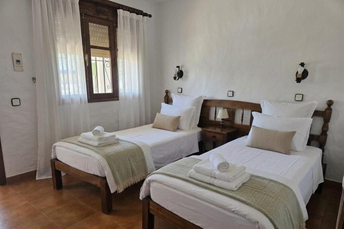 A hotel room with two single beds, a window with curtains, a wooden headboard, and bedside lamps.