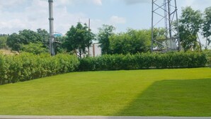 A well-maintained grassy area with a metal fence, trees, and industrial structures in the background. - Regenta Place The Golden Castle Baddi (Baddi)