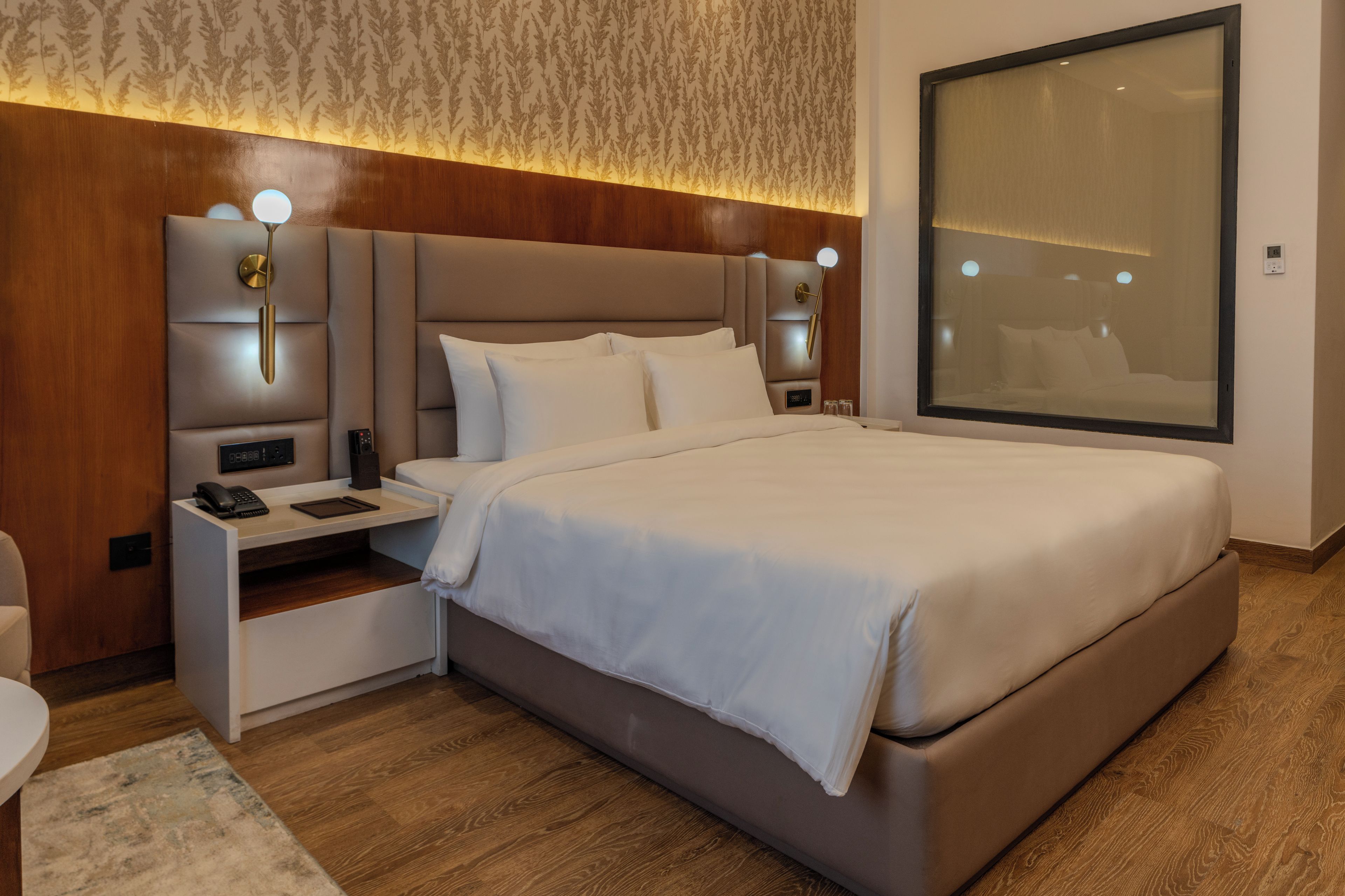 Quarto de hotel moderno com uma cama grande, mesas de cabeceira, luminária na parede e uma janela ampla.