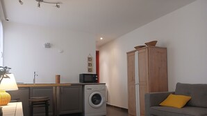 A compact living space with a kitchenette, a washing machine, a grey sofa with a yellow pillow, and a wooden wardrobe. - Fully-equipped T2 apartment in a country setting close to all amenities, 15 minutes from the beach. (Solliès-Ville)