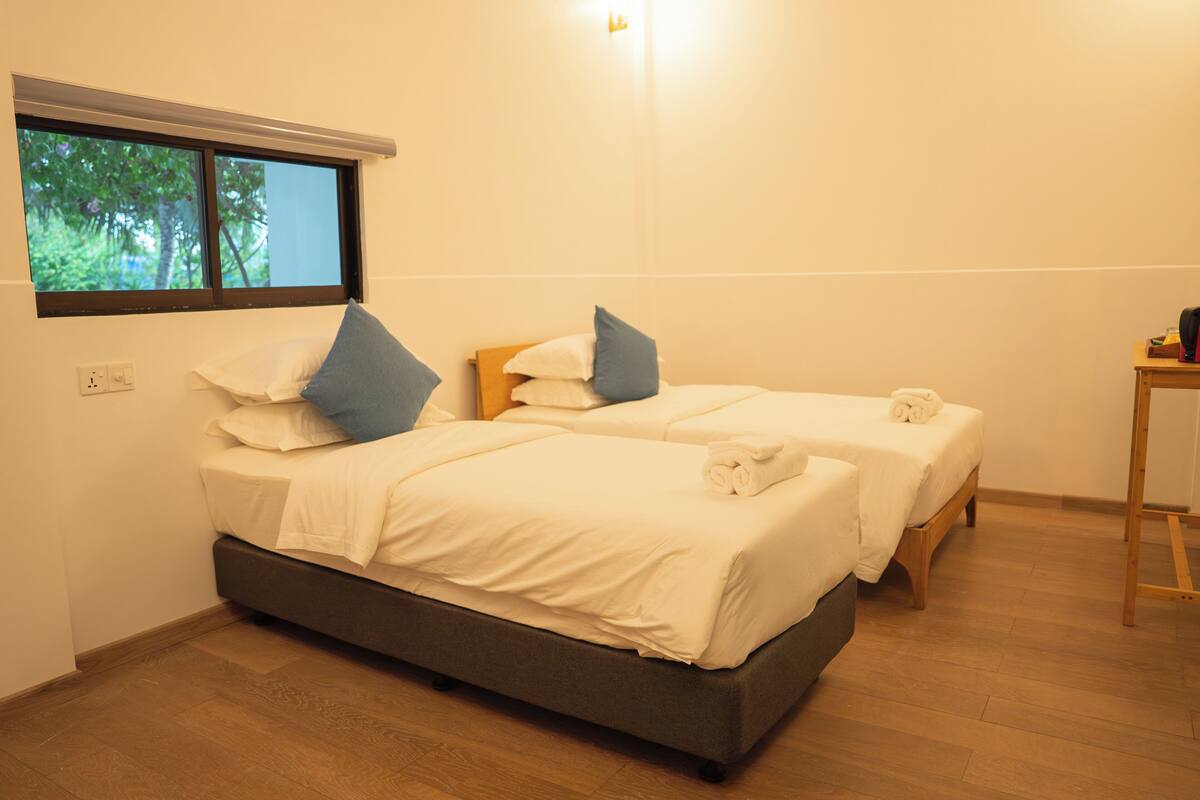 A room with two beds, a window with a view of trees, a wooden floor, and a small table.