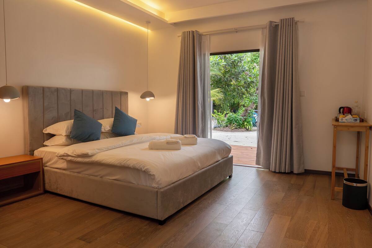 A modern bedroom with a large bed, bedside lamps, a wooden nightstand, a small table, and a view of greenery outside.