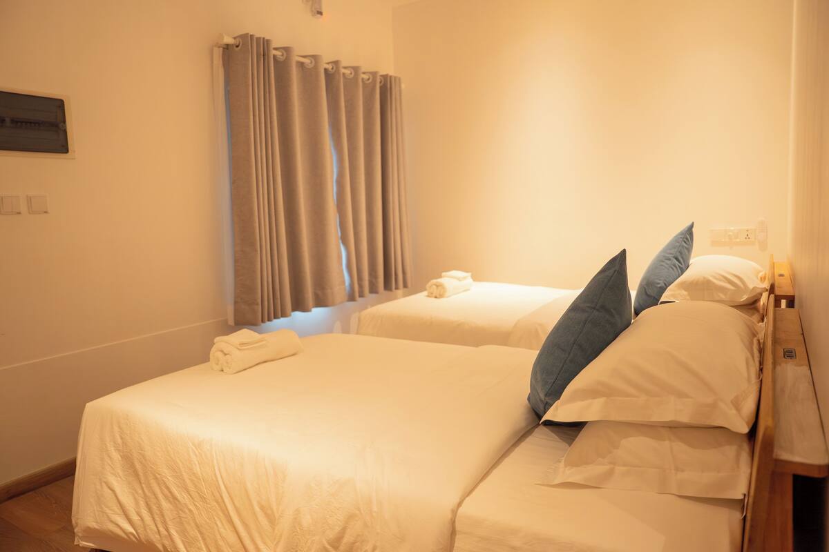 A hotel room with two beds, a window with curtains, a wall-mounted air conditioning unit, and a bedside table.