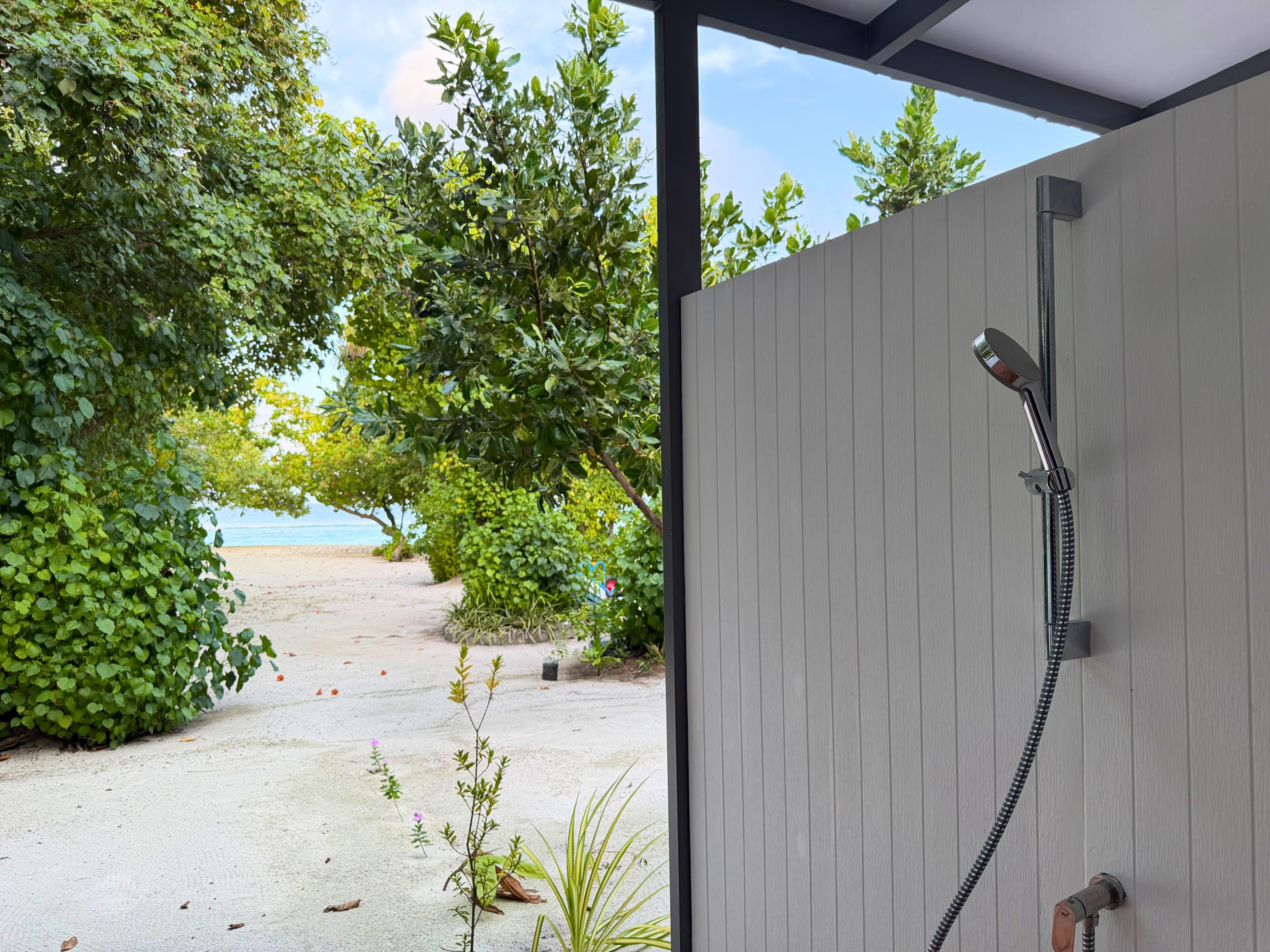 A covered outdoor area with a shower, surrounded by greenery and a view of the beach.