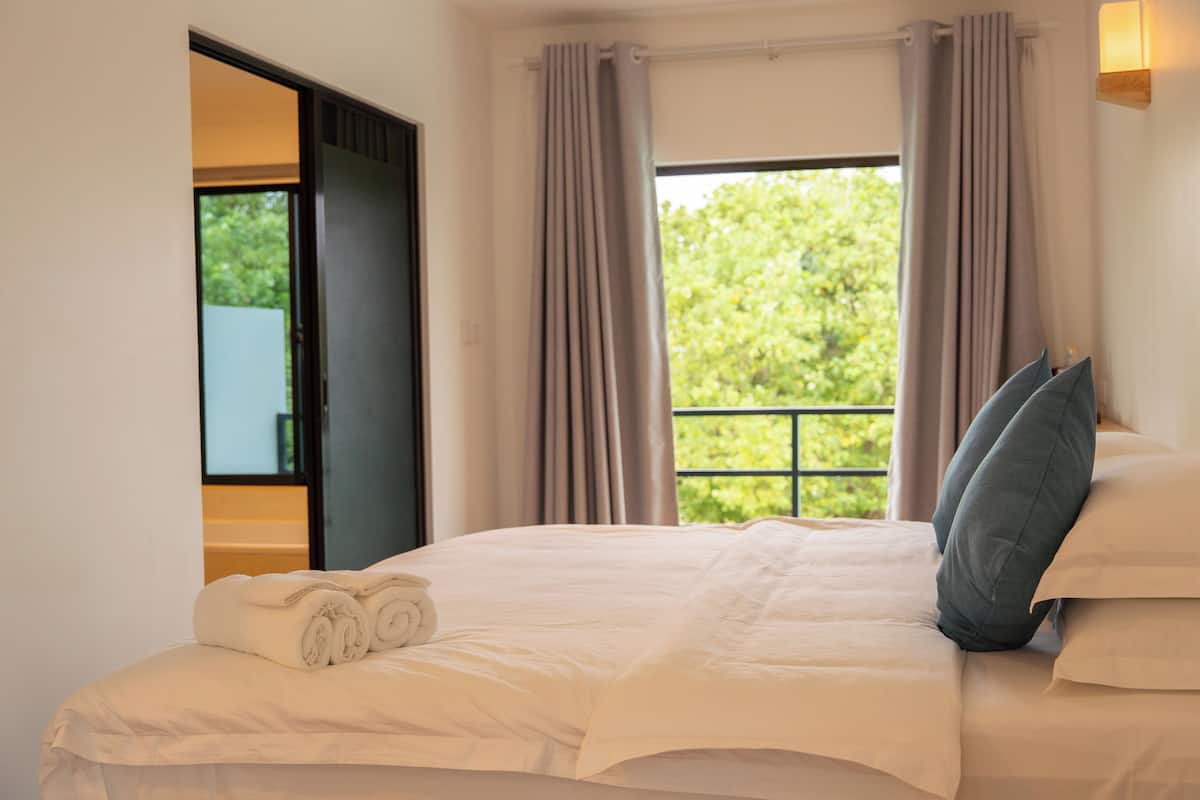 A hotel room with a bed, a window with curtains, a view of greenery, and a door leading to an outdoor area.