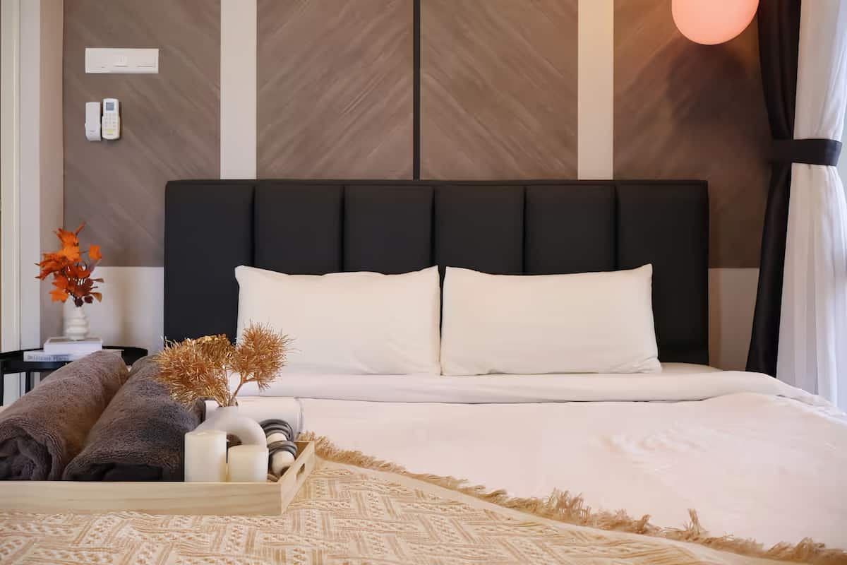 A neatly made bed with white linens, a dark headboard, and a bedside table with a vase of dried flowers.