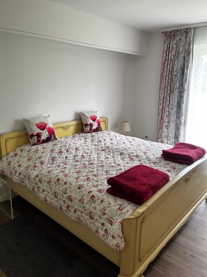 A bed with a floral quilt, two pillows, a lamp, and a window with curtains. - First floor apartment 3 bedrooms rural location (Malente)