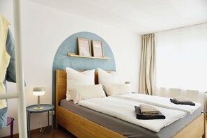Een tweepersoonsbed met houten frame, een rond nachtkastje met lamp, een hoofdbord met een blauw boogvormig element en een raam met gordijnen.