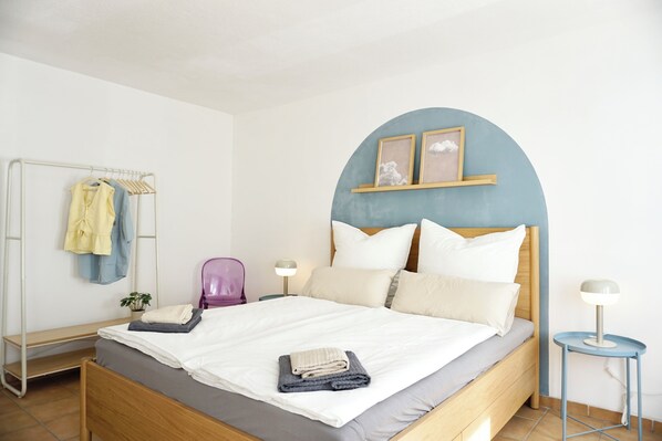 Een slaapkamer met een bed, nachtkastje, lamp, stoel, kast en een plank met foto’s.