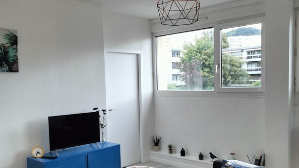 A modern living room with a sofa, a television on a blue cabinet, a round coffee table, and a window with a view of buildings and trees. - Apparently T2 45 m2, Vallières district  (Clermont-Ferrand)