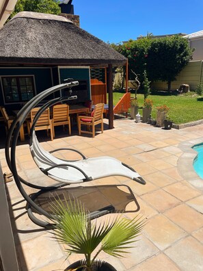 Eine Terrasse mit Pool, einem strohgedeckten Gebäude und einem Holztisch mit Stühlen.