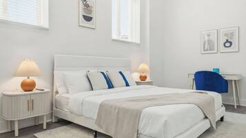 A modern bedroom with a white bed, bedside lamps, a desk, and artwork on the walls.