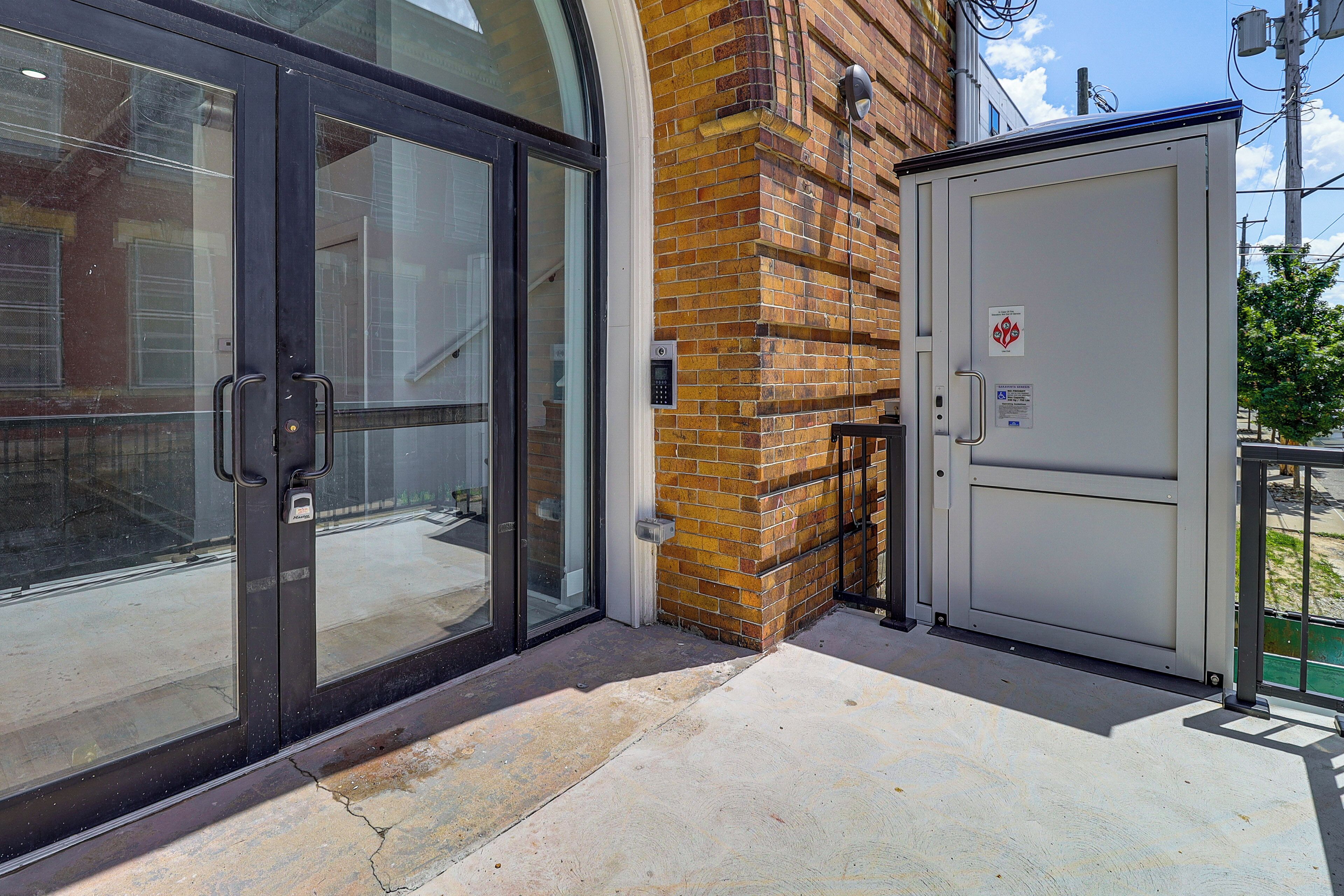Une entrée d’immeuble avec des portes en verre, un mur en briques et un coffret électrique.