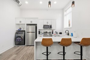 A modern kitchen with white cabinets, a built-in microwave, a refrigerator, and a washing machine. There are three bar stools with brown seats.