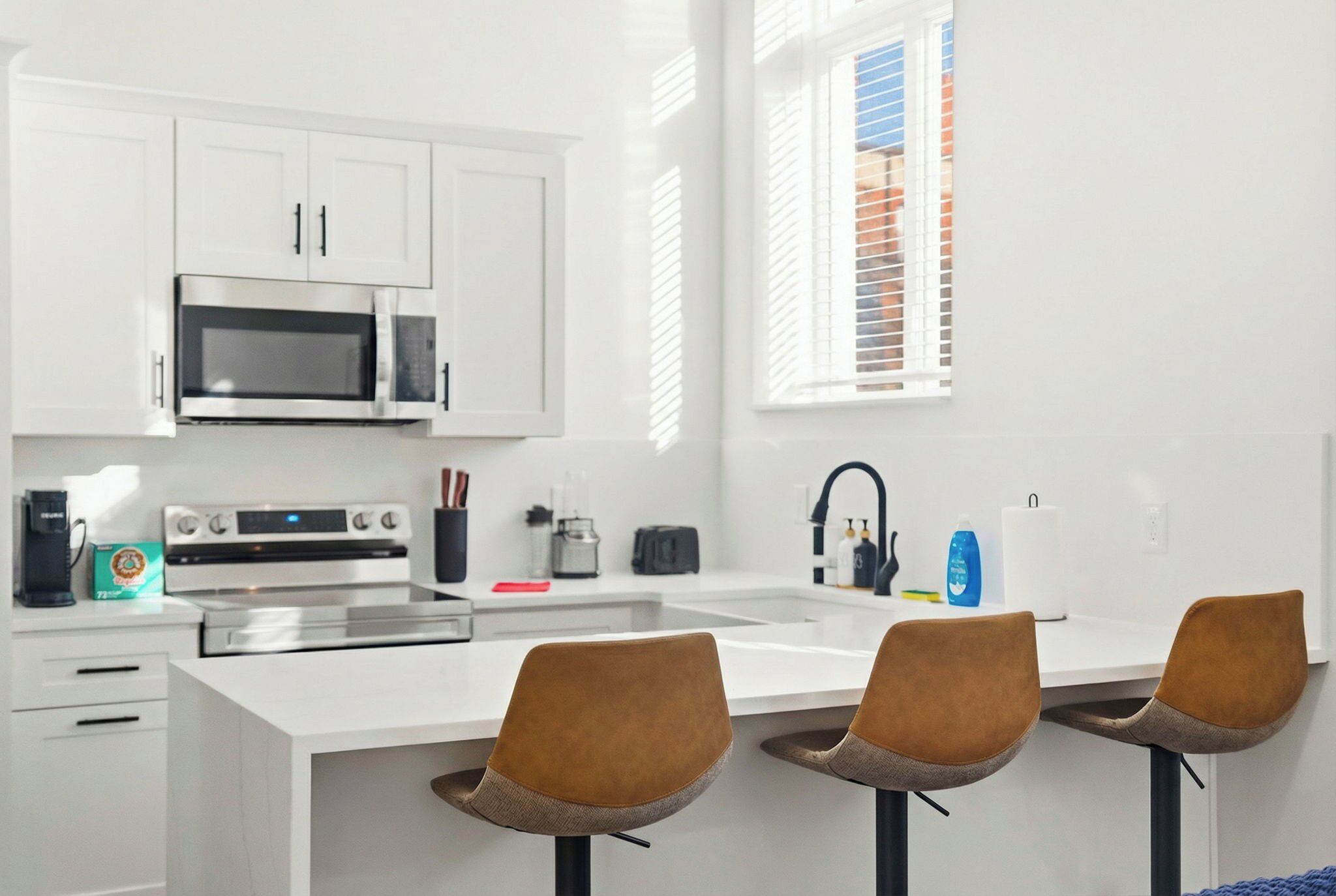 A modern kitchen with white cabinetry, a microwave, an oven, and a sink. There are three brown chairs at a white table.