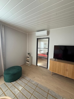 Una sala de estar moderna con una televisión de pantalla plana, un reposapiés verde, un mueble de madera y un ventanal corredizo que da a un balcón.