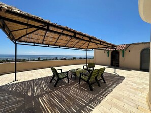 Ein  überdachter Außenbereich zum Sitzen mit Blick auf das Meer, einem Tisch und Stühlen.