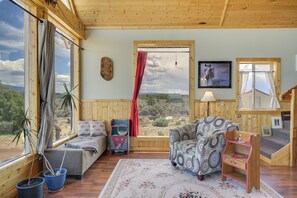 A cozy room with a bed, a patterned armchair, a small table, and a window with a view of the landscape. - Sangre de Cristo Views! Retreat Near Wild Horses! (San Luis)